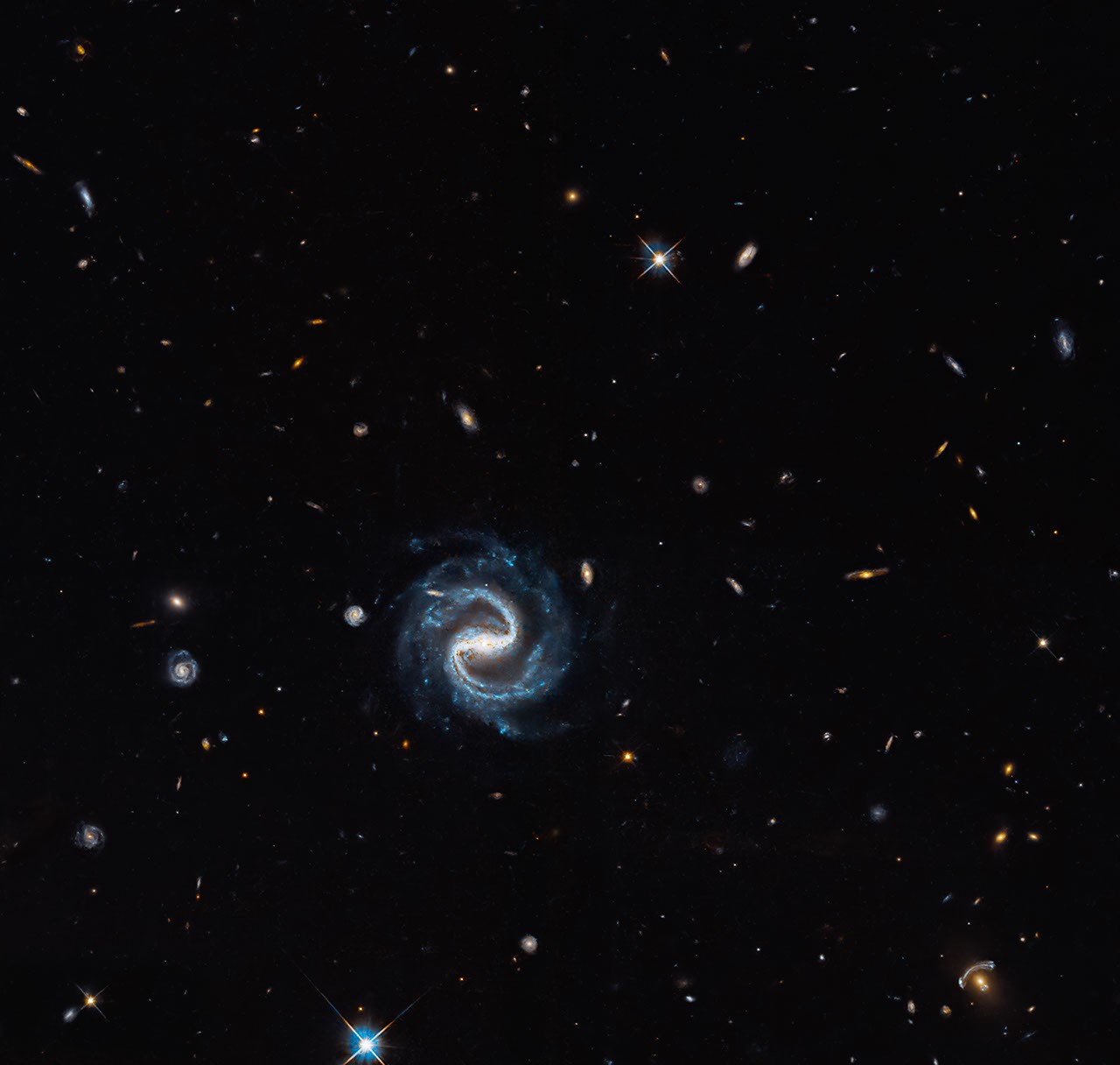 Hubble Space Telescope view showing a foreground elliptical galaxy gravitationally lensing a distant background galaxy into curved blue arcs beside a barred spiral galaxy.