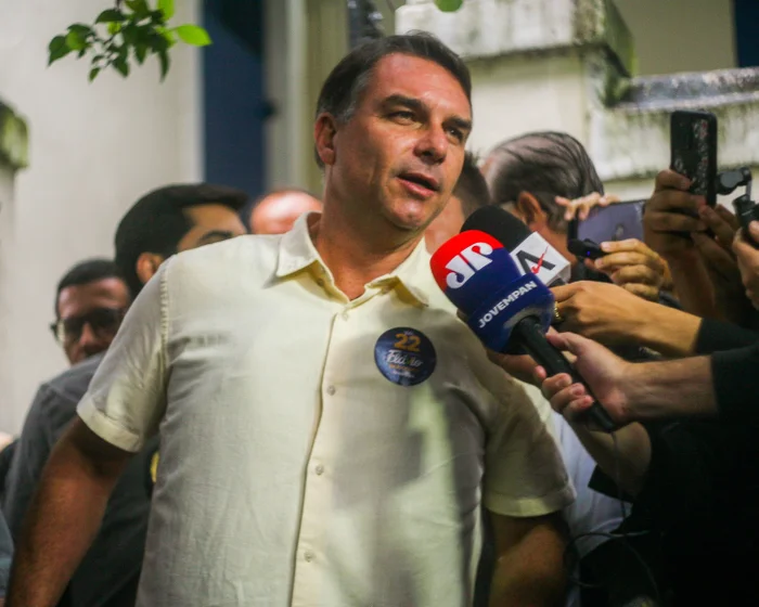 Flávio Bolsonaro speaks to reporters holding microphones.